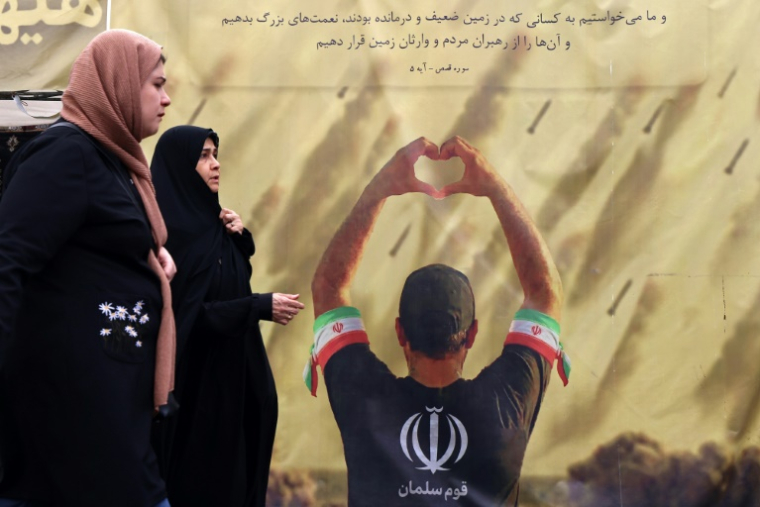 Des femmes passent devant une banderole représentant un homme faisant un cœur avec ses mains devant des tirs de missiles, à Téhéran le 19 avril 2026 ( AFP / ATTA KENARE )