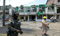 Un membre du Groupe des opérations spéciales de la police nationale colombienne monte la garde tandis que des agents de nettoyage déblayent les débris du site d'une explosion à la bombe survenue la veille à Cali, en Colombie, le 22 août 2025 ( AFP / Iusef Samir Rojas )