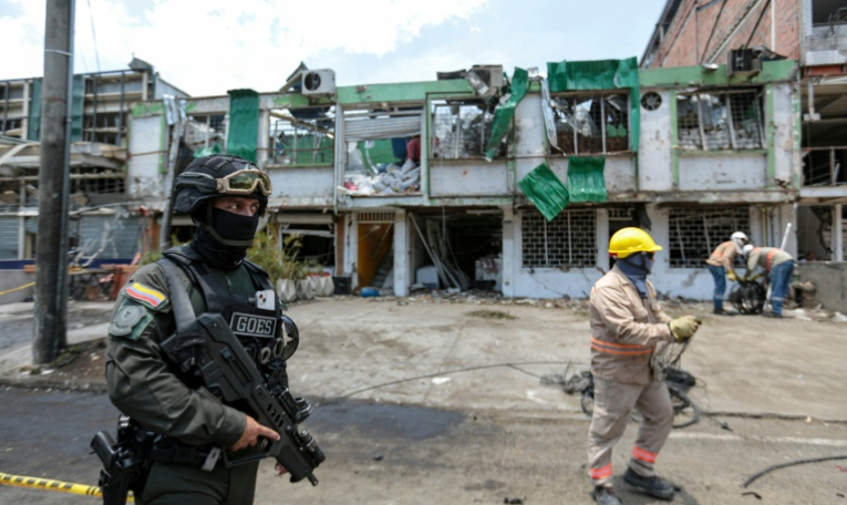 Un membre du Groupe des opérations spéciales de la police nationale colombienne monte la garde tandis que des agents de nettoyage déblayent les débris du site d'une explosion à la bombe survenue la veille à Cali, en Colombie, le 22 août 2025 ( AFP / Iusef Samir Rojas )