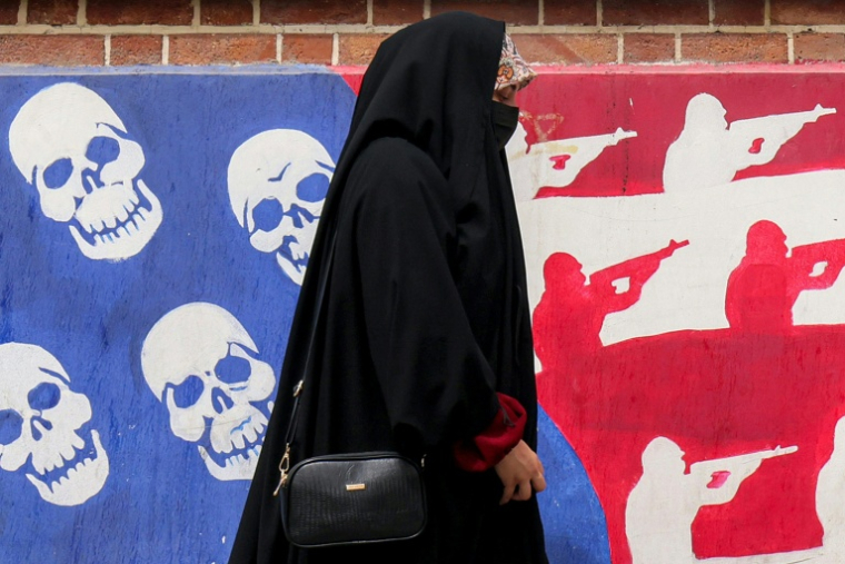 Une femme passe devant un mur de l'ancienne ambassade des Etats-Unis à Téhéran, peint aux couleurs du drapeau américain avec les étoiles remplacées par des têtes de mort et les rayures par des silhouettes de soldats, le 8 avril 2025 en Iran ( AFP / ATTA KENARE )