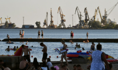 Des estivants profitent de la plage à Marioupol, citée ukrainienne contrôlée par la Russie, avec le port de commerce à l'arrière-plan, le 14 juillet 2025 ( AFP / Olga MALTSEVA )