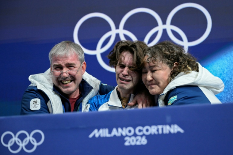 Le Kazakhstanais Mikhail Shaidorov (au centre) avec ses entraîneurs au moment où sa note s'affiche après son programme libre de patinage artistique le 13 février 2026 où il a décroché l'or à la surprise générale ( AFP / WANG Zhao )