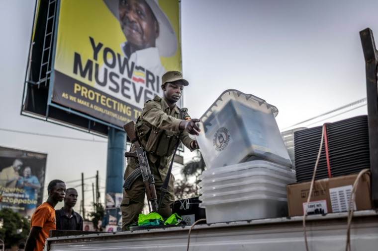 Un policier décharge d'un camion des urnes destinés à un bureau de vote devant une affiche électorale du président sortant Yoweri Museveni, candidat du Mouvement de résistance nationale (NRM) à la présidentielle, le 15 janvier 2026 à Kampala, en Ouganda ( AFP / Luis TATO )