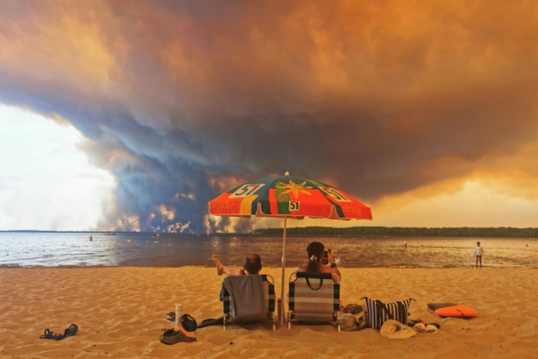Des vacanciers sont assis sur la plage du Moutchic, à Lacanau, dans le sud-ouest de la France, le 24 juillet 2026, tandis que d'épais nuages de fumée s'élèvent dans le ciel au-dessus d'un incendie de forêt ( AFP / Maximilien LAMY )