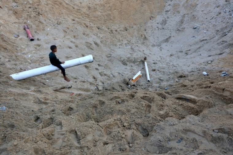 Un enfant palestinien est assis sur un bout d'une conduite d'eau dans un immense cratère provoqué par un raid israélien à Gaza-ville, le 28 juin 2025 ( AFP / Omar AL-QATTAA )
