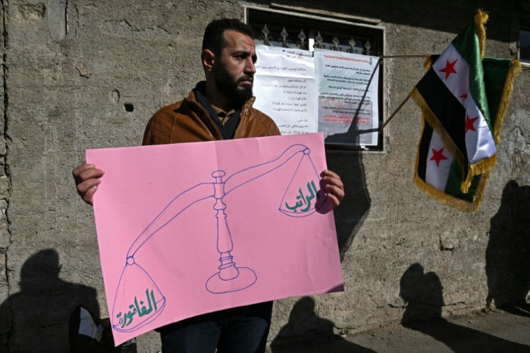 Un homme tient une pancarte lors d'une manifestation contre la hausse des prix de l'électricité en Syrie, à Damas le 29 janvier 2026, montrant d'un côté de la balance "le salaire" et de l'autre, à gauche, "la facture" ( AFP / LOUAI BESHARA )