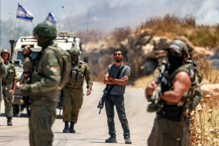 Un colon israélien se tient entre des soldats israéliens rassemblées devant des Palestiniens le long d'une route qui s'étaient rassemblés pour protester contre des colons israéliens qui auraient mis le feu à une zone du village palestinien d'Idna, à l'ouest d'Hébron en Cisjordanie occupée, le 5 juin 2026 ( AFP / HAZEM BADER )