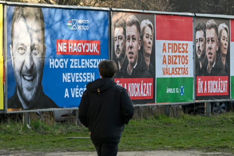 Un piéton devant des panneaux d'affichage gouvernementaux montrant le président ukrainien Volodymyr Zelensky (à gauche), avec le texte "Ne laissons pas Zelensky avoir le dernier mot", et une autre affiche montrant la présidente de la Commission européenne Ursula von der Leyen (à droite), le président ukrainien Volodymyr Zelensky (au centre) et le chef de l'opposition hongroise Peter Magyar (à gauche) avec le texte "Ils sont eux-mêmes le risque", à Budapest le 3 mars 2026 ( AFP / Attila KISBENEDEK )