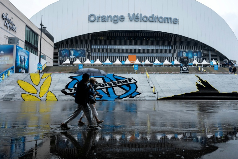 Des piétons abrités sous un parapluie passent devant le stade Vélodrome, où le match de L1 OM-PSG a été reporté de 24 heures en raison des intempéries, le 21 septembre 2025 à Marseille ( AFP / Miguel MEDINA )