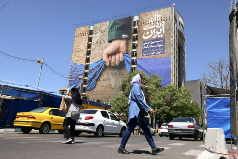 Des passants près d'un panneau d'affichage sur la façade d'un immeuble représentant le détroit d'Ormuz avec une inscription en persan disant "À jamais dans la main de l'Iran", place Vanak à Téhéran, le 25 mai 2026 ( AFP / ATTA KENARE )