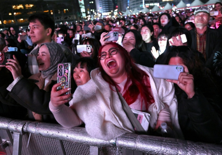 Des fans de BTS assistent au concert de retour du boys band de K-pop devant un écran installé sur le lieu de l'événement à Séoul, le 21 mars 2026 ( AFP / Jung Yeon-je )