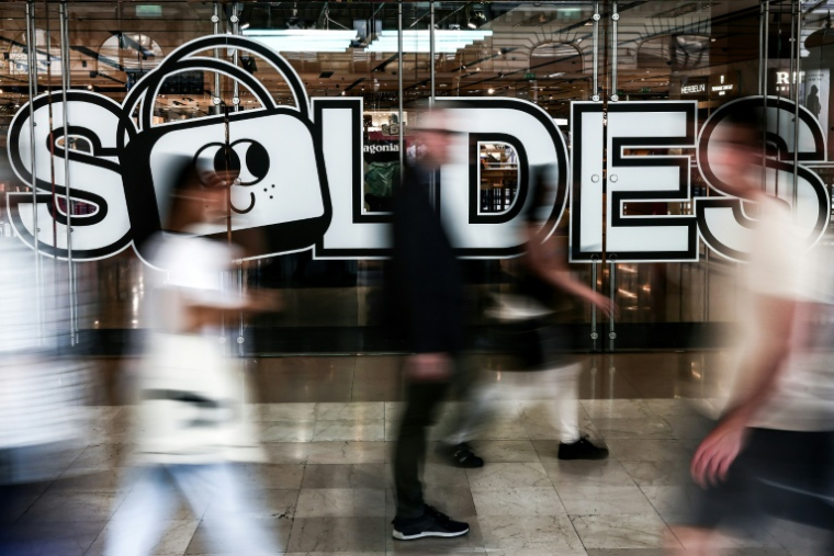 Des passants passent devant la porte d'entrée d'un grand magasin où est accrochée une affiche annonçant les soldes d'été à Paris, le 25 juin 2025 ( AFP / Thibaud MORITZ )