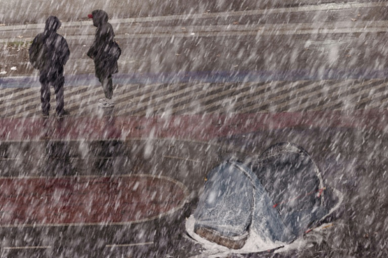 Two pedestrians stand next to a homeless person's tent during a snowfall in Paris on January 5, 2026. ( AFP / JOEL SAGET )