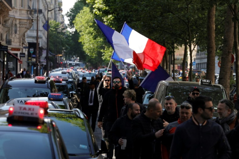 Des chauffeurs de taxi brandissent un drapeau français lors d'un rassemblement pour protester contre les nouvelles conditions tarifaires du transport sanitaires et la concurrence des VTC, à Paris, le 19 mai 2025 ( AFP / GEOFFROY VAN DER HASSELT )