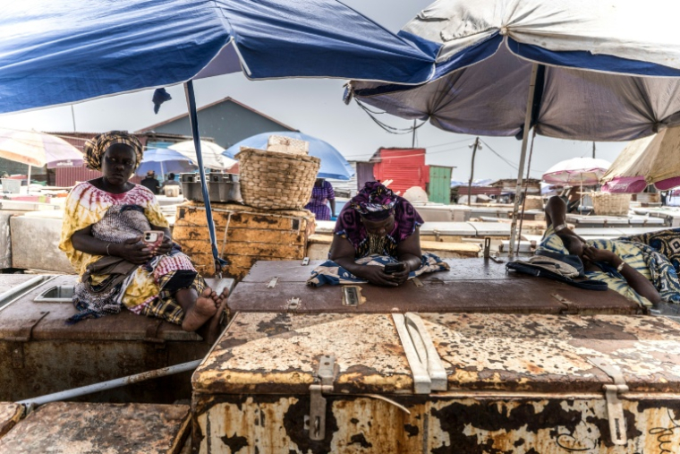 Des poissonniers se reposent sur leurs réfrigérateurs au point de débarquement des poissons à Tanji, en Gambie, le 12 septembre 2026, alors que les coupures d'électricité prolongées rendent difficile la conservation de leurs prises ( AFP / MUHAMADOU BITTAYE )