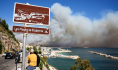 la plage des Corbières, à Marseille, dans le sud de la France, le 8 juillet 2025, des passants observent la fumée d'un incendie de forêt qui fait rage en arrière-plan ( AFP / Clement MAHOUDEAU )