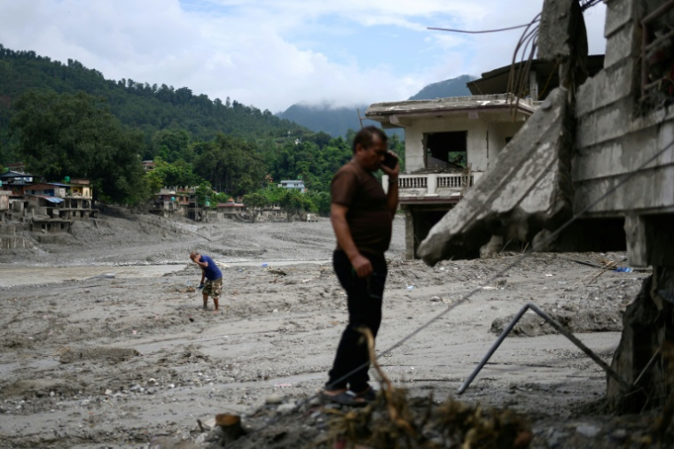 Deux hommes au téléphone au milieu d'habitations recouvertes de boue et de débris après la crue destructrice qui a fait plus de 600 morts à la frontière entre le Népal et la Chine, le 29 août 2026 à Devighat, dans le district népalais  de Nuwakot  ( AFP / Manan VATSYAYANA )
