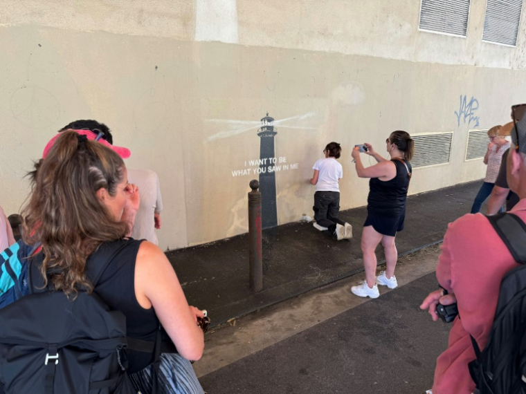 Des passants prennent des photos pendant que la peintre en décor du patrimoine, Agnès Perrone, répare une œuvre d'art de l'artiste de rue Banksy représentant un phare sur la façade d'un bâtiment, après qu'elle a été vandalisée pendant la nuit à Marseille, le 1er juin 2025 ( AFP / Viken KANTARCI )