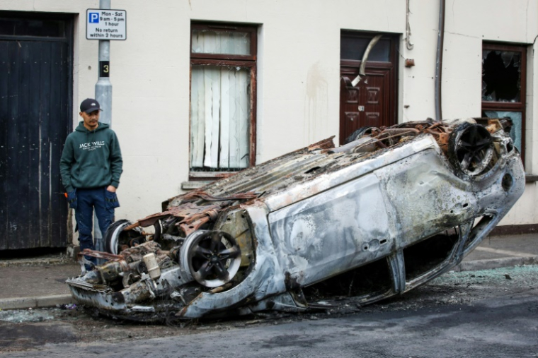 Un passant observe les restes d'une voiture calcinée, au lendemain d'une nouvelle nuit d'émeutes visant des immigrés, le 11 juin 2025 à Ballymena, en Irlande du Nord ( AFP / PAUL FAITH )