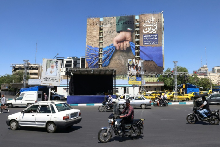 Des véhicules devant un panneau d'affichage sur la façade d'un bâtiment représentant le détroit d'Ormuz avec une légende en persan disant "À jamais dans la main de l'Iran", place Vanak à Téhéran, le 25 mai 2026 ( AFP / ATTA KENARE )