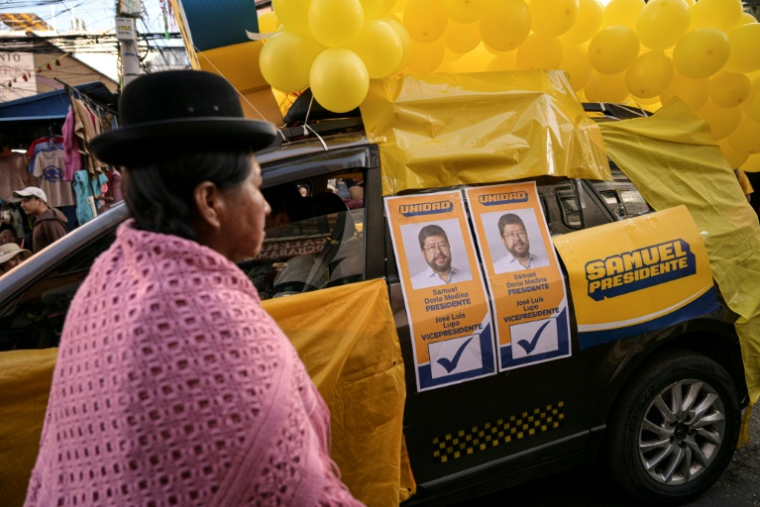 Une femme autochtone bolivienne passe devant une voiture avec des affiches de campagne du candidat présidentiel de la coalition Alianza Unidad, Samuel Doria Medina, à La Paz le 12 août 2025. ( AFP / MARTIN BERNETTI )