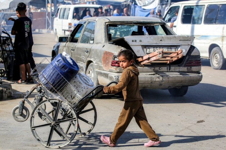 Une fillette palestinienne pousse un fauteuil roulant cassé chargé de jerrycans le long d'une route dans un camp de déplacés à Nousseirat, dans le centre de la bande de Gaza, le 7 octobre 2025 ( AFP / BASHAR TALEB )