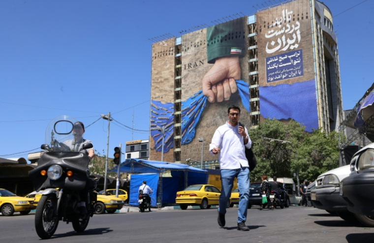 Des passants traversent une rue devant un panneau d'affichage apposé sur la façade d'un immeuble représentant le détroit d'Ormuz, avec une légende en persan indiquant "Pour toujours entre les mains de l'Iran", à Téhéran, le 25 mai 2026 ( AFP / ATTA KENARE )