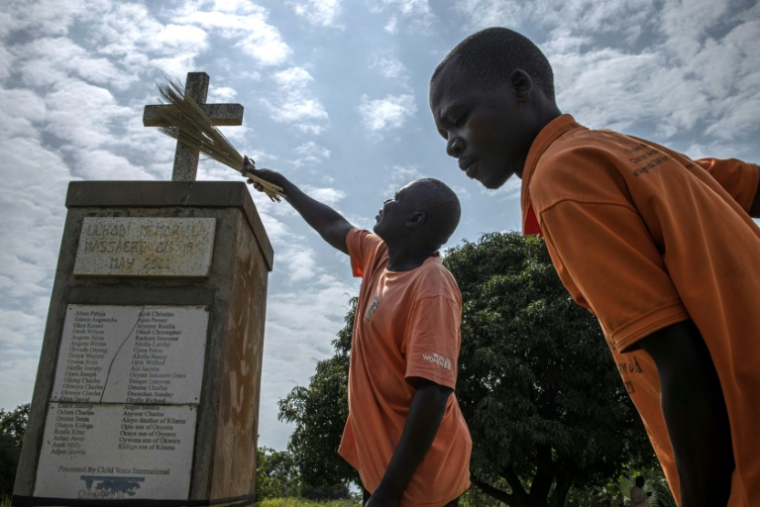 Wilfred Lalobo (centre), 60 ans, un survivant du massacre de Lukodi le 19 mai 2004, et Richard Ochola, 34 ans, un employé du mémorial de Lukodi, nettoie une stèle commémorative du massacre perpétré il a plus de 20 ans pzr la guérilla de la LRA dans ce village du nord de l'Ouganda, le 4 septembre 2025. ( AFP / Stuart Tibaweswa )