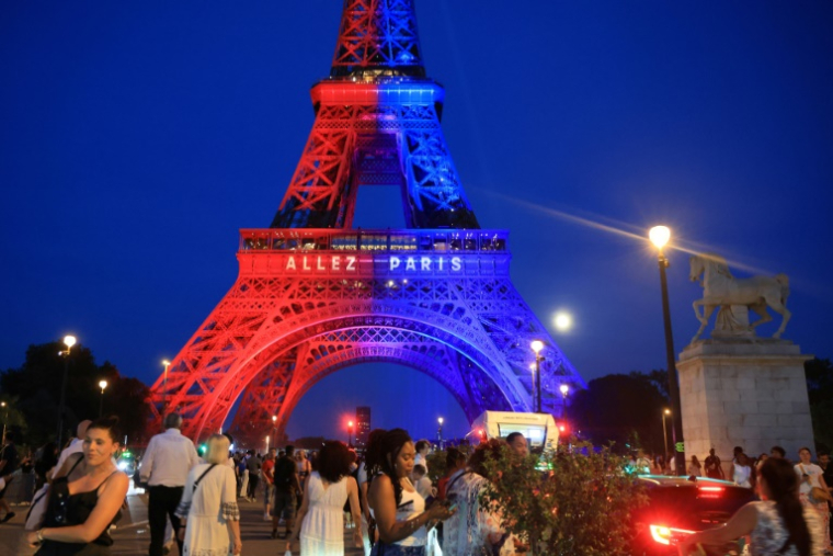 Des passants déambulent sous la Tour Eiffel, illuminée aux couleurs du Paris Saint-Germain (PSG) et sur laquelle est inscti le message "Allez Paris", avant la finale de la Ligue des champions de l'UEFA opposant le PSG à Arsenal, le 29 mai 2026 à Paris ( AFP / Ludovic MARIN )