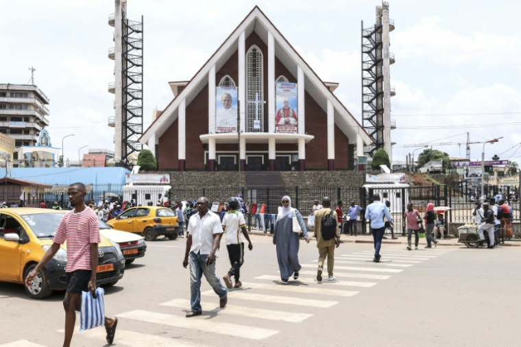Des piétons marchent dans une rue près de la cathédrale Notre-Dame-des-Victoires, où sont exposés des portraits du pape Léon XIV, à Yaoundé, le 8 avril 2026 ( AFP / Daniel BELOUMOU OLOMO )