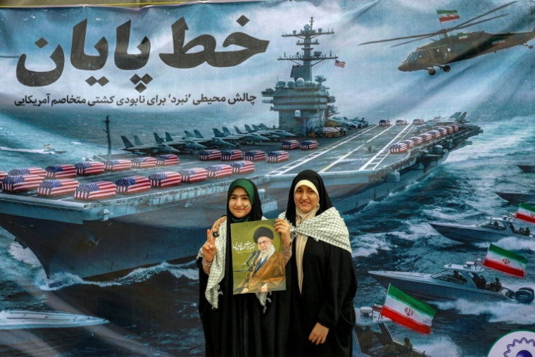 Des femmes posent avec un portrait du guide suprême iranien, Ali Khamenei, devant une affiche montrant des cercueils entassés sur le pont d'un porte-avions américain, lors d'un défilé pour le 47e anniversaire de la Révolution islamique à Téhéran, le 11 février 2026 ( AFP / - )