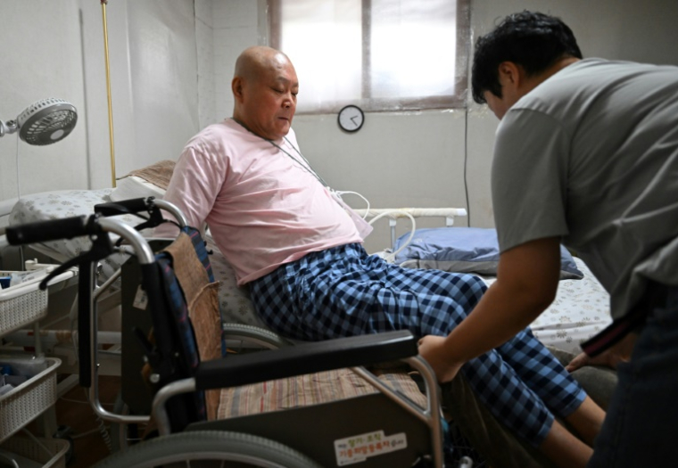 Lee Myung-shik, en train de se mettre au lit avec l'aide d'un soignant, chez lui, sur l'île de Jeju, en Corée du Sud, le 9 juin 2026 ( AFP / Jung Yeon-je )