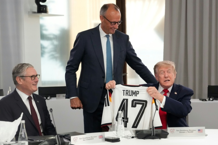Le président américain Donald Trump (d) montre le maillot de football allemand floqué à son nom offert par le chancelier allemand Friedrich Merz (c) en présence du Premier ministre britannique Keir Starmer, le 16 juin 2026 au sommet du G7 à Evian ( POOL / Thibault Camus )