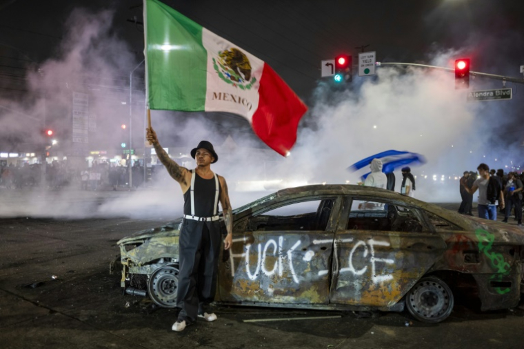 Un manifestant tient un drapeau mexicain devant l'épave d'une voiture incendiée lors de heurts avec les forces de l'ordre contre l'expulsion d'immigrés, à Los Angeles, le 7 juin 2025 en Californie ( AFP / RINGO CHIU )