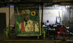 (ARCHIVES) Des habitants regardent la télévision à côté d'une affiche représentant le président guinéen Mamadi Doumbouya avant l'annonce des résultats provisoires officiels du référendum à Conakry, le 23 septembre 2025. ( AFP / PATRICK MEINHARDT )