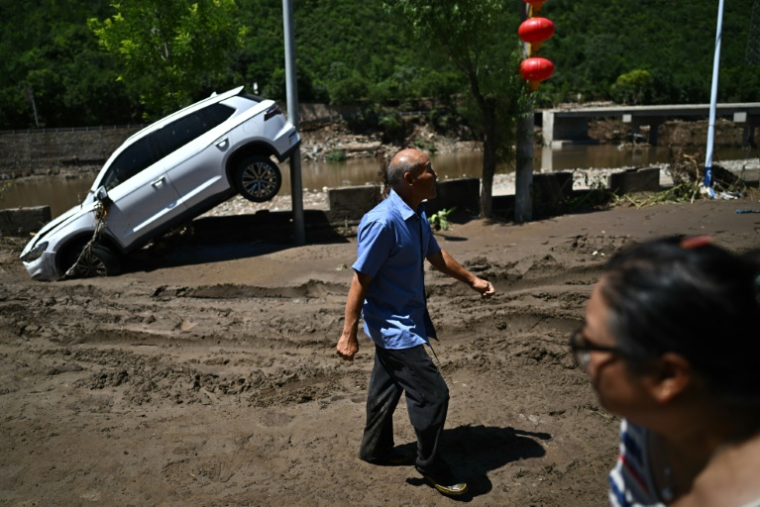 Des personnes passent devant une voiture endommagée par les inondations dans le district de Huairou, à la périphérie de Pékin, le 30 juillet 2025 ( AFP / Pedro Pardo )