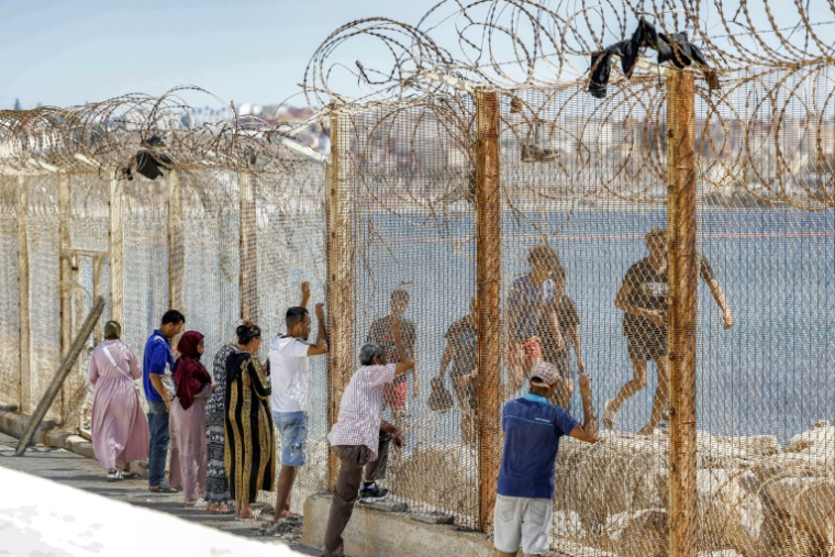 Des proches de migrants ayant traversé la frontière entre le Maroc et l'enclave espagnole de Ceuta voient passer des personnes refoulées derrière un grillage, dans la ville marocaine de Fnideq, le 2 août 2026 ( AFP / Abdel Majid BZIOUAT )