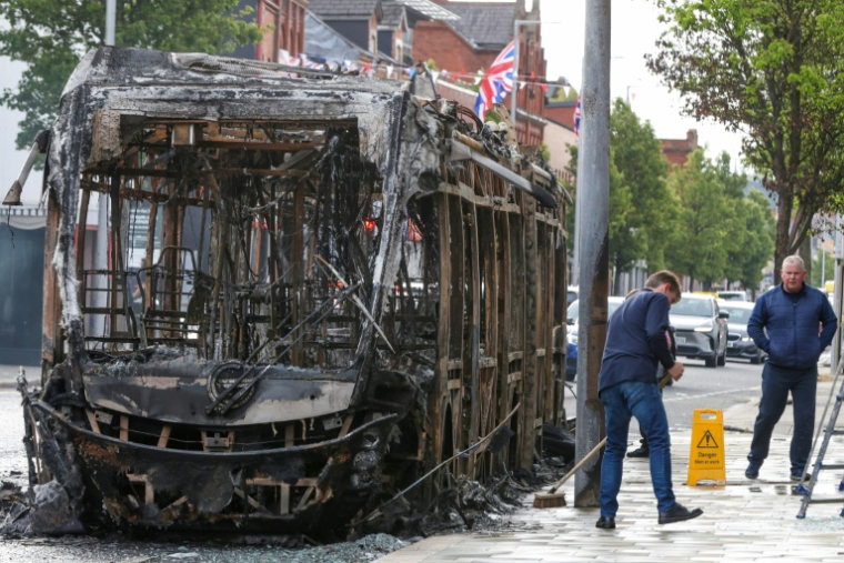 Nettoyage de la rue à côté d'un bus calciné après des manifestations qui ont dégénéré en violences la nuit précédente, dans l'est de Belfast, en Irlande du Nord, le 10 juin 2026 ( AFP / Paul Faith )