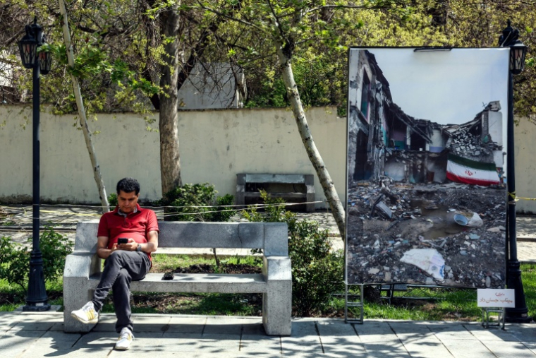 Un Iranien consulte son téléphone, assis sur un banc à côté d'une photo montrant "la guerre au Moyen-Orient", le 16 avril 2026 dans un parc de Téhéran ( AFP / - )