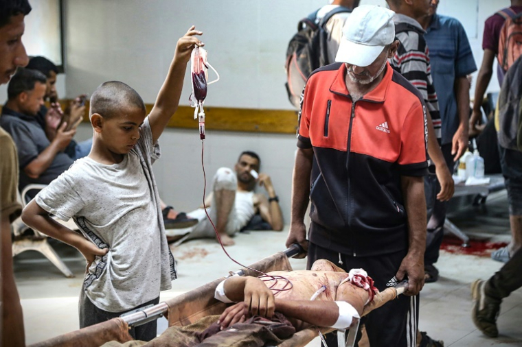 Un garçon palestinien tient une poche de sang pour un homme blessé à un point de distribution d'aide dans le secteur d’Al-Tina, à Khan Younès, dans le sud de la bande de Gaza, à l’hôpital Nasser, le 19 juillet 2025 ( AFP / - )