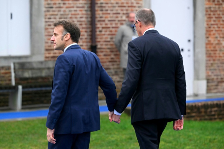 Emmanuel Macron (G) et Friedrich Merz se tiennent la main à leur arrivée au sommet informel de l'UE au château belge d'Alden Biesen en Belgique le 12 février 2026 ( AFP / NICOLAS TUCAT )
