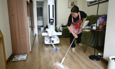 Un robot assistant ménager transporte une bouteille jusqu'à une poubelle tandis qu'une femme de ménage nettoie le sol chez une cliente à Pékin, le 21 mai 2026 en Chine ( AFP / WANG Zhao )