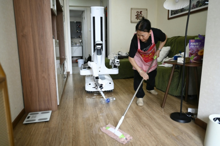 Un robot assistant ménager transporte une bouteille jusqu'à une poubelle tandis qu'une femme de ménage nettoie le sol chez une cliente à Pékin, le 21 mai 2026 en Chine ( AFP / WANG Zhao )