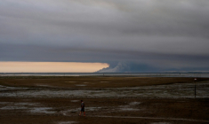 Un homme marche sur une plage d'Andernos au bord du bassin d'Arcachon, au fond duquel s'élèvent les fumées de l'incendie de Biscarosse, le 23 juillet 2026 ( AFP / Olivier MORIN )