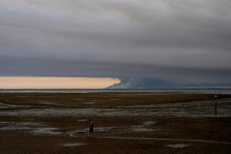Un homme marche sur une plage d'Andernos au bord du bassin d'Arcachon, au fond duquel s'élèvent les fumées de l'incendie de Biscarosse, le 23 juillet 2026 ( AFP / Olivier MORIN )