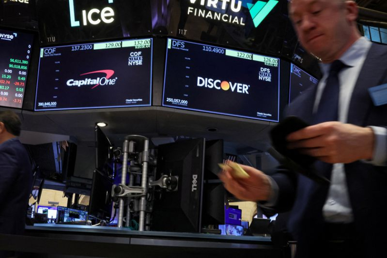 Des écrans affichent les logos et les informations sur les transactions de Capital One Financial et de Discover Financial pendant que les traders travaillent sur le parquet de la Bourse de New York