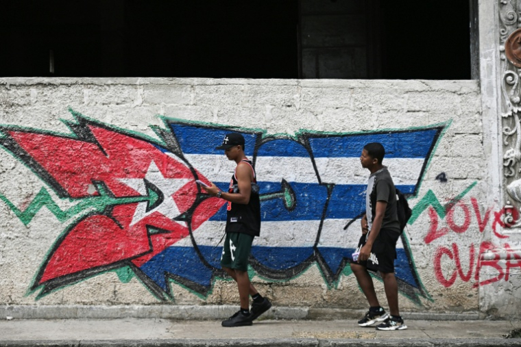 TDeux jeunes passent devant un mur arborant un drapeau cubain, le 14 juillet 2026 à La Havane, lors d'une panne d'électricité nationale à Cuba ( AFP / YAMIL LAGE )