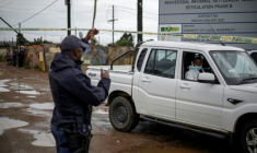 Un policier sud-africain soulève la bande jaune barrant l'accès pour permettre à un expert de la police scientifique d'approcher la scène d'une tuerie dans un bar clandestin, le 21 décembre 2025 à Bekkersdal (Afrique du Sud) ( AFP / EMMANUEL CROSET )