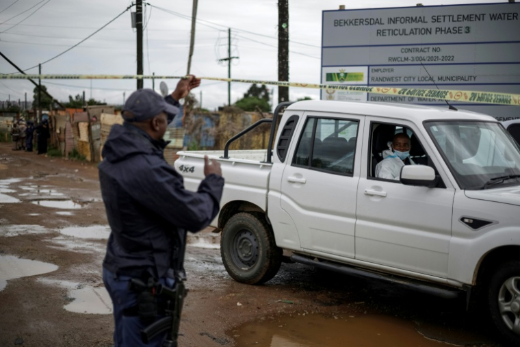 Un policier sud-africain soulève la bande jaune barrant l'accès pour permettre à un expert de la police scientifique d'approcher la scène d'une tuerie dans un bar clandestin, le 21 décembre 2025 à Bekkersdal (Afrique du Sud) ( AFP / EMMANUEL CROSET )