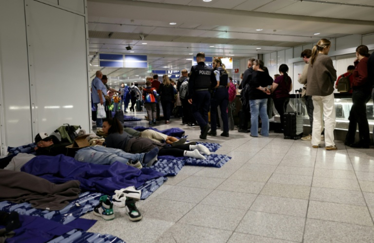 Des passagers dorment sur des matelas gonflables posés à même le sol tandis que d'autres font la queue à côté d'eux à l'aéroport international de Munich, dans le sud de l'Allemagne, tôt le matin du 4 octobre 2025 ( AFP / Michaela STACHE )